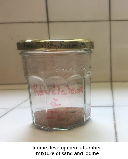 Picture of a jam jar containing a brown powder. The jar has the handwritten mention : developer at I2. The photo is captioned: diiodine development tank: mixture of sand and diiodine.