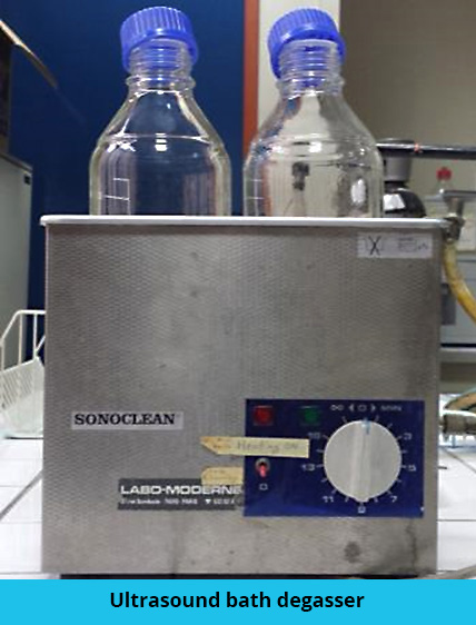 Photo of an ultrasonic degasser. It is a metal tank with an adjustment wheel on its front face. Two half-opened bottles are inside the tank.
